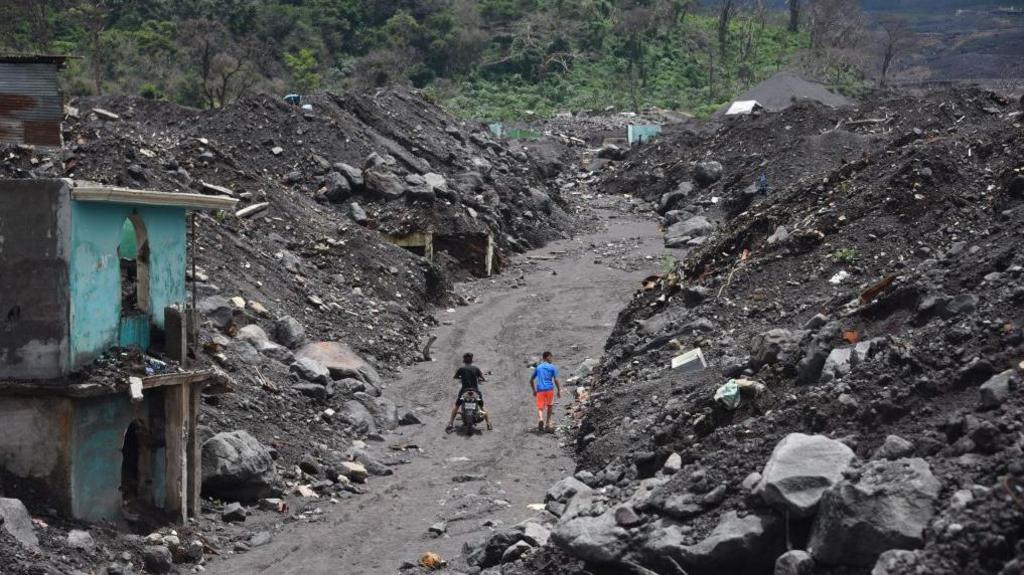 A street carved through huge piles of rock and ash, with a few ruined houses