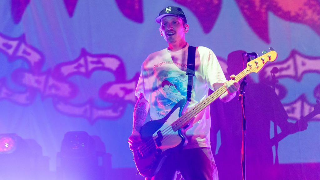 Seb from pop-punk band Neck Deep is wearing a black cap, a whte t-shirt and holds a black bass guitar. He is stood on a stage with purple lighting.