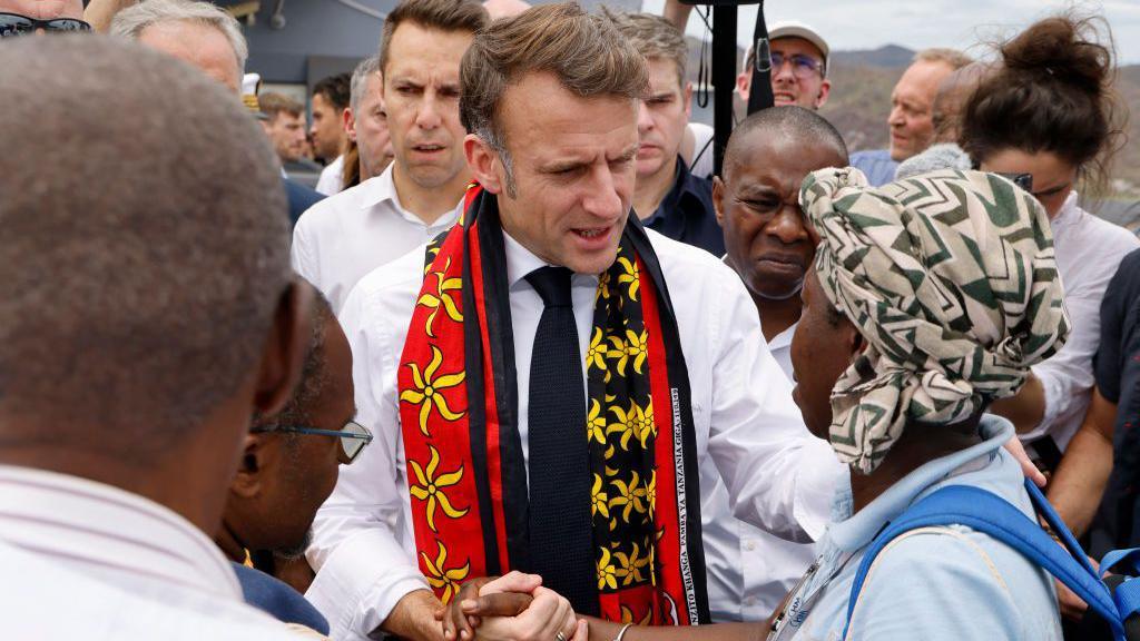 French President Emmanuel Macron, wearing a bright red and yellow scarf, holds hand of a woman in Mayotte with crowd behind them  