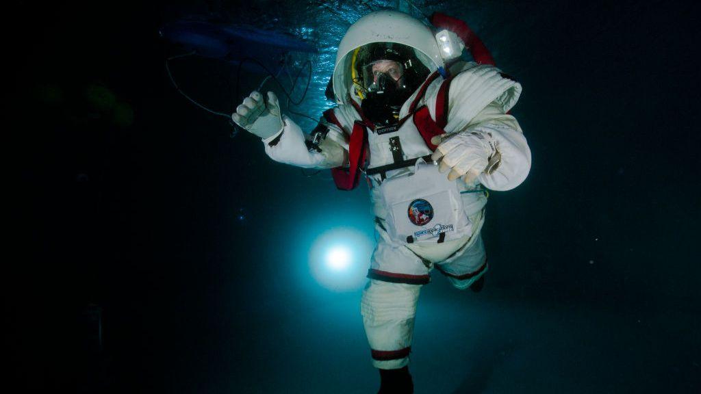 A woman wearing a white spacesuit taking part in underwater training at Nasa. The bulky white suit is surrounded in darkness but for a single light illuminating a small area of the pool.