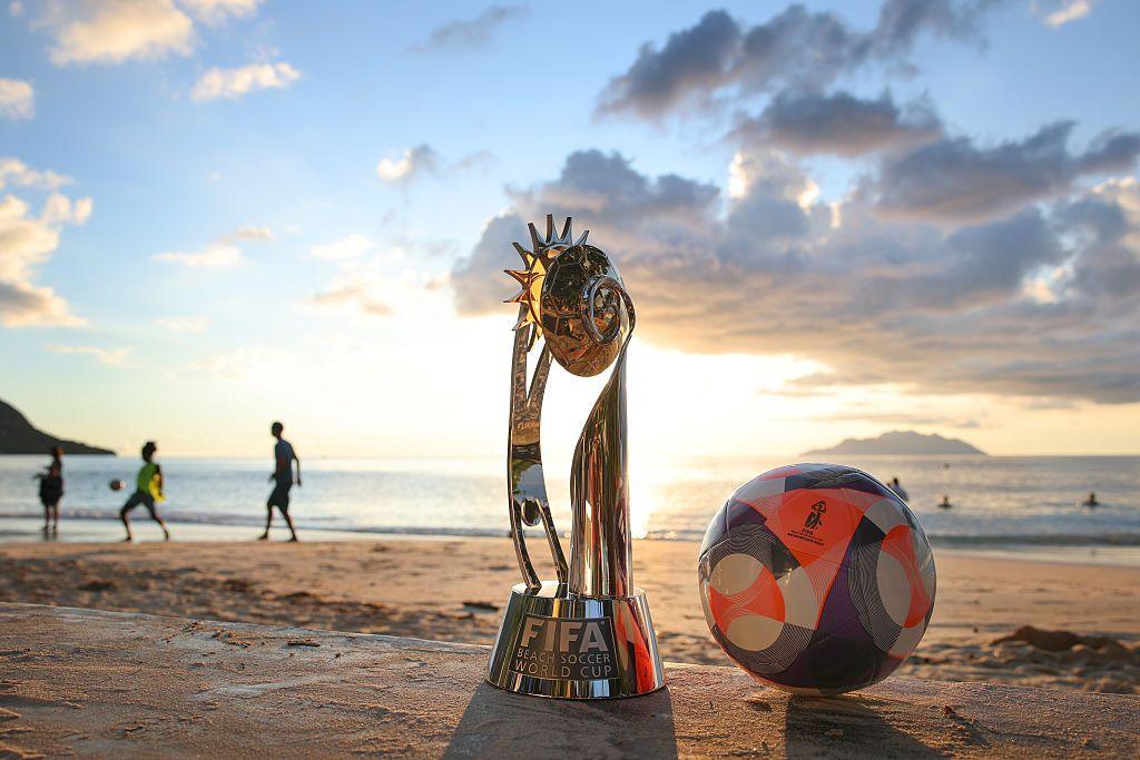 A detailed view of the trophy and the official match ball as people play football at Beau Vallon beach. The sun is low is the sky above the sea in the background.