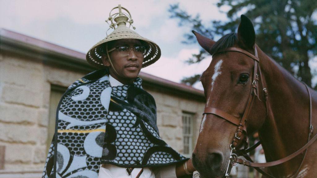 Moshoeshoe II wears glasses, a traditional blanket and mokorotlo . He holds a horse by its reins.