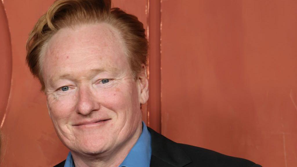 Conan O'Brien, in a blue shirt and dark suit coat, stands in front of an orange backdrop as he attends the HBO & Max Post-Emmy Reception at San Vicente Bungalows