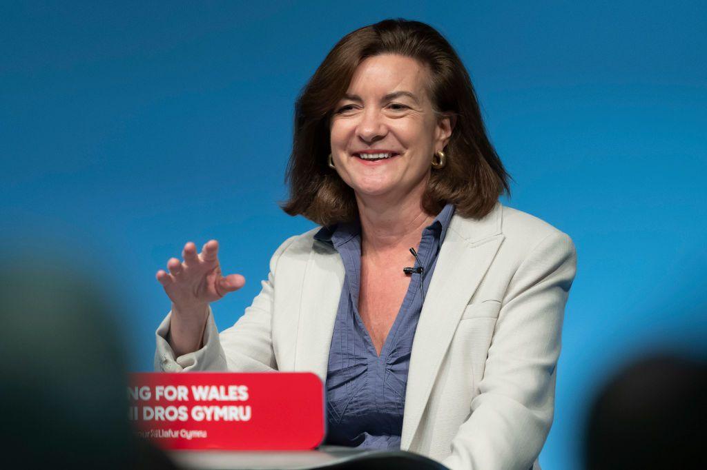 Eluned Morgan smiling as she gives her speech to the Welsh Labour conference. She is standing behind a podium with red sign with the words "Delivering for Wales - Welsh Labour" on it in white lettering, but the words are partly obscured by the head of someone in the audience. She is wearing a light jacket and dark blue blouse