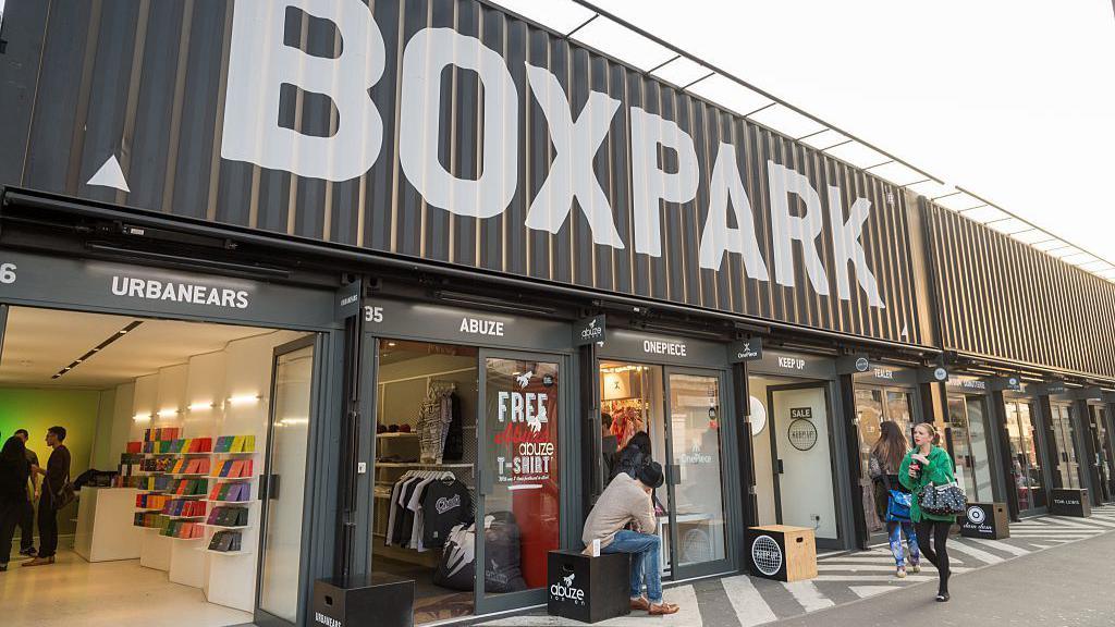 We can see several containers are lined up next to each other with shops inside them. About them is a big sign saying BOXPARK, which is printed on corrugated metal.