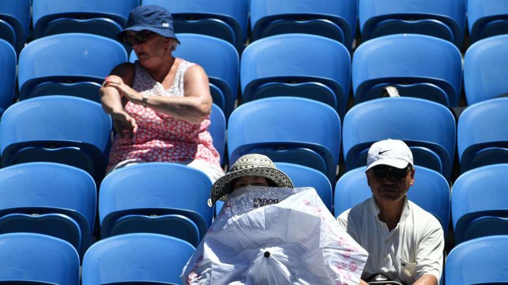 A spectator uses an umbrella to shield herself from the sun as another applies sunscreen during the men's singles third round match between Luxembourg's Gilles Muller and Spain's Pablo Carreno Busta on day five of the Australian Open tennis tournament in Melbourne on January 19, 2018.