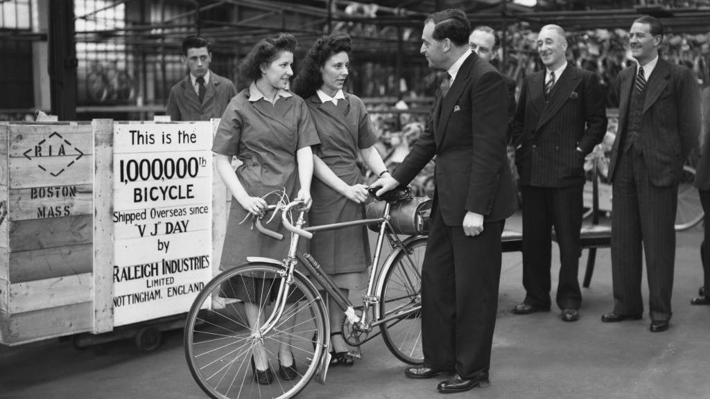 A black and white photograph of a politician talking to two women who are holding a bicycle in front of a sign announcing this is the 1,000,000th bicycle made since the war ended 