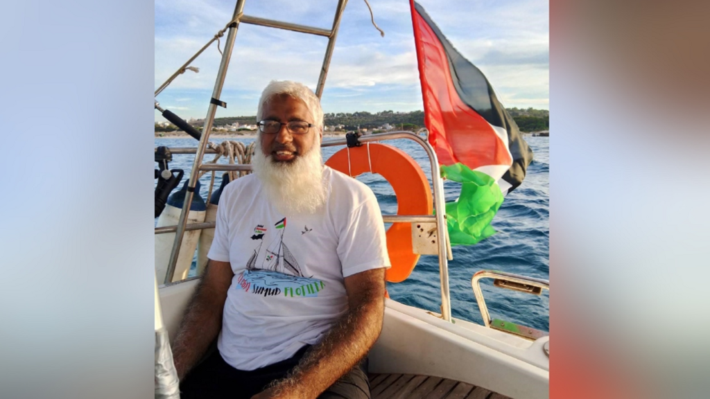 Mr Hussain is wearing a T'shirt and shorts and is sat on the boat at sea. His is sitting on wooden slats with a rubber ring and Palestinian flag behind him with the expanse of the sea visible and boat paraphernalia to one side. 
