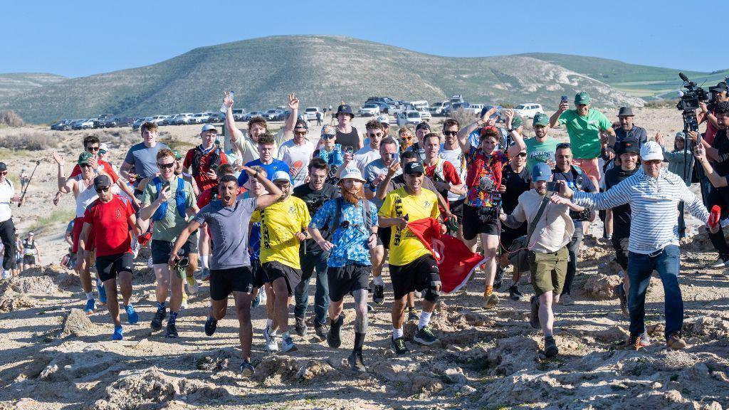 Russ Cook surrounded by running supporters as he crosses a finish line in Tunisia. He is wearing a blue England football shirt and a bucket hat. 