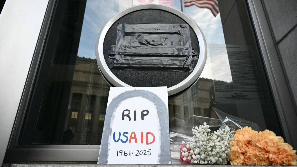 Black tape covers a USAID sign outside it's headquarters in Washington. A sign with a grave stone reads "RIP USAID". Flowers sit nearby 