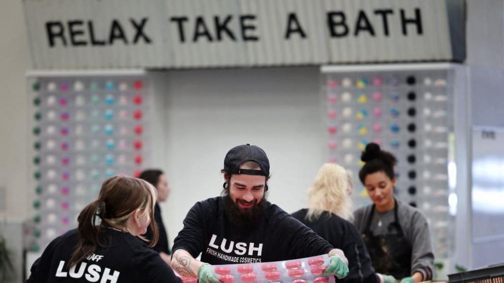 A group of men and women making colourful bath bombs in moulds. They are wearing black t-shirts that say Lush. There is a sign on the wall that says Relax Take a Bath.