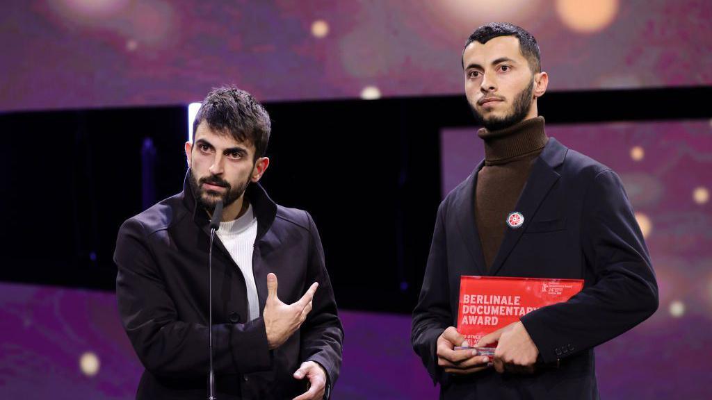 Yuval Abraham stands at a microphone and speaks as he gestures towards Basel, holding an award, on stage