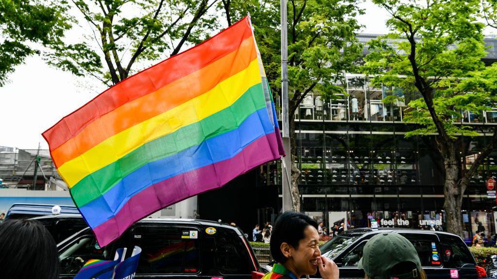 A large rainbow flag - a symbol of LGBTQ rights - flying in the wind, attached to a pole.
