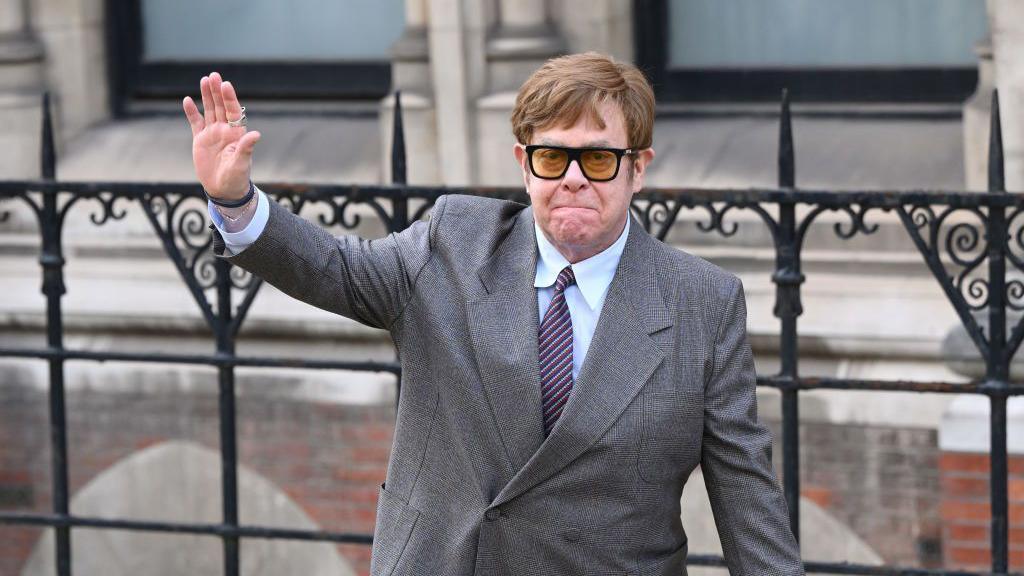 Sir Elton John in a suit, tie and sunglasses, waving outside the Royal Courts of Justice in 2023
