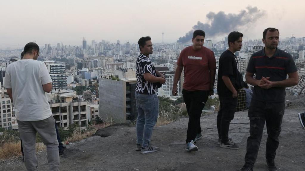 Five men stand on a hill overlooking Tehran. Behind where four of them are gathered on the right, dark smoke rises from a strike on the city