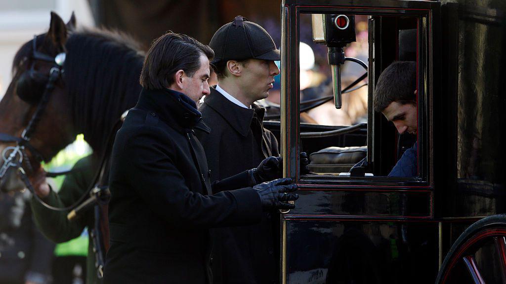 Benedict Cumberbatch dressed as Sherlock Holmes getting into a horse-drawn carriage, with two other men, both in black, around him