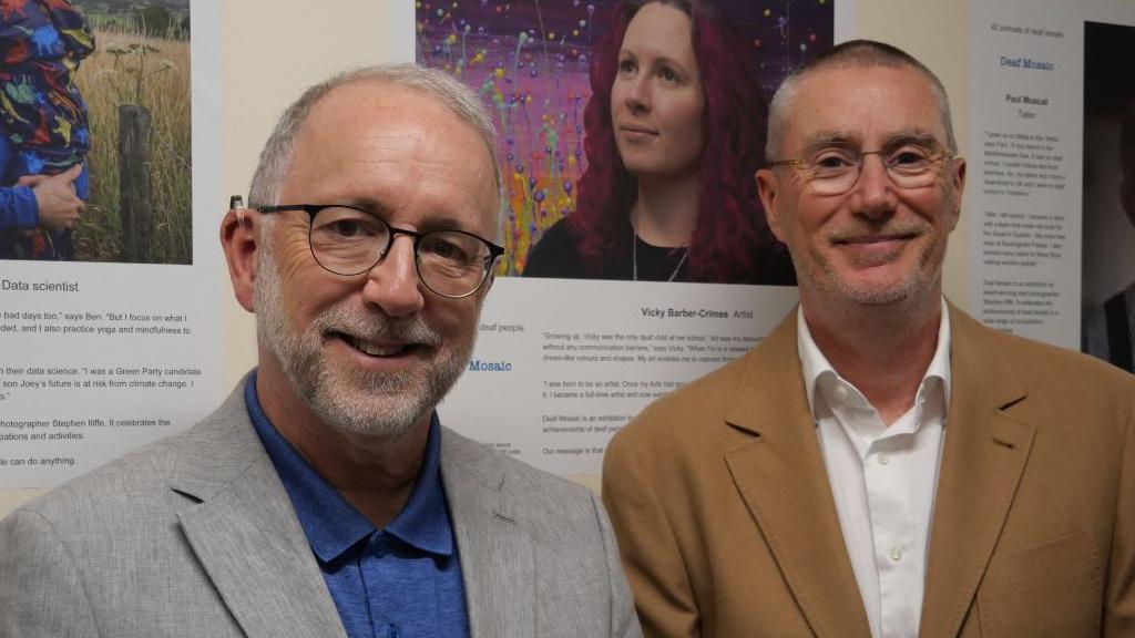 Chief Executive Officer at Nottingham University Hospitals, Anthony May stands alongside photography Stephen Iliffe standing in front of the exhibiton. Both men are wearing glasses, Anthony on the left is in a grey suit blazer while Stephen has a brown suit jacket on. 