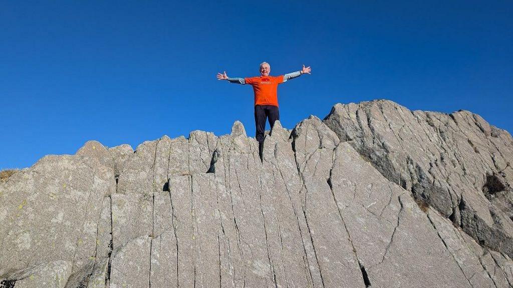 Ian stands on top of a rocky mountain. The sky is blue. He has his arms outstretched and wears an orange T-shirt with the words Maggie's on it, which is the name of the charity he is raising money for. He looks very happy and somewhat excited.