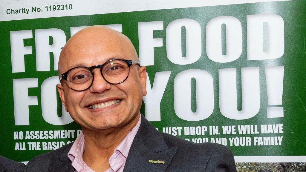 A man smiling at the camera in front of a banner. He is wearing a grey suit with a pink shirt and black glasses. The banner is green with the words "free food for you! in white letters.