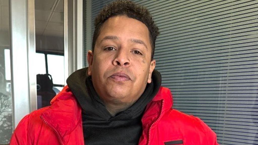 A man with black curly hair, a black top and bright red coat in front of a window with blinds drawn