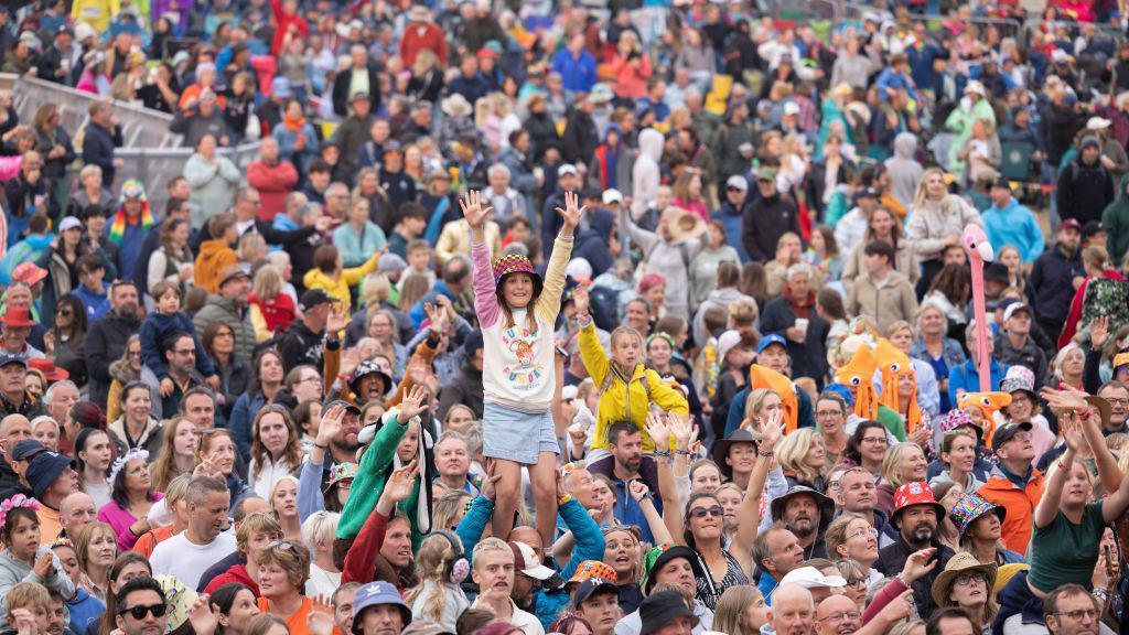 A large, cheerful crowd at a festival, with a young girl on someone's shoulders raising her arms in excitement.