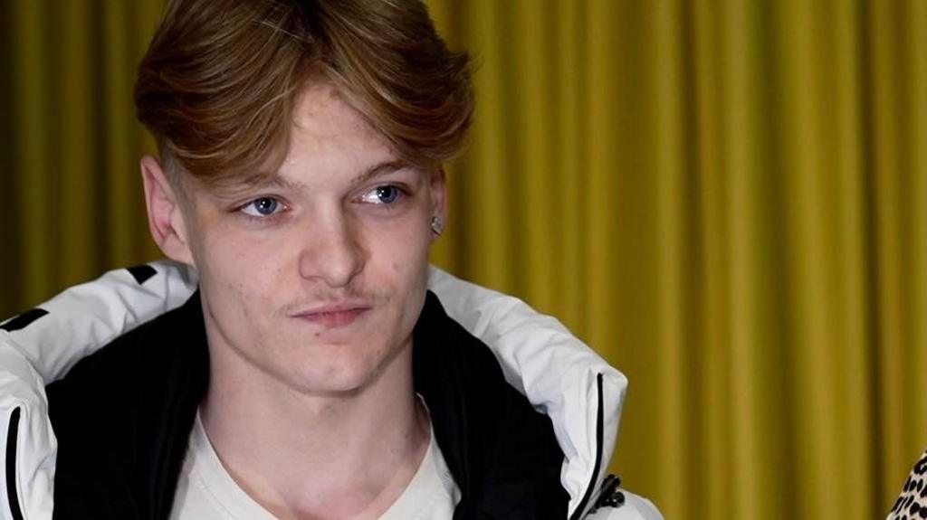 Mason Read, who is wearing a white coat with a collar around his neck and a white T-shirt. He has light brown hair in curtains and is looking to the right of the camera with a slight pout. There are yellow curtains behind him.