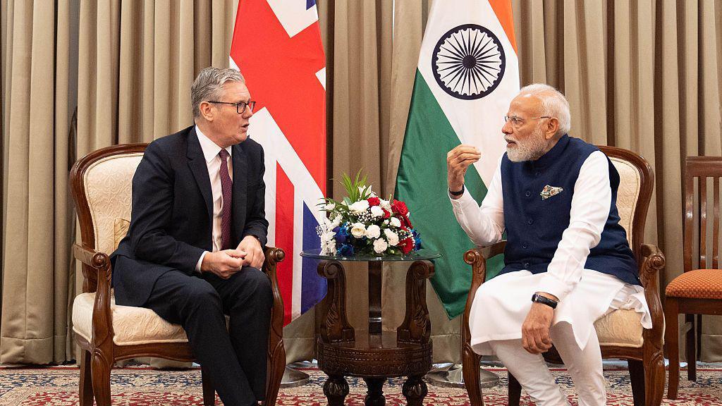 UK Prime Minister Sir Keir Starmer and Indian Prime Minister Narendra Modi sat in cream coloured chairs, in conversation. They are sat in front of large flags of their respective nations and an arrangement of white, blue and red flowers is on a small table between them.