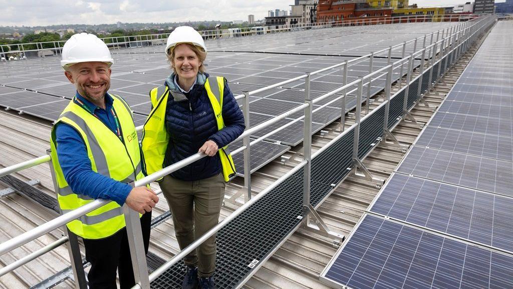 two people in high vis jackets stand on an elevated metal walkway on top of a roof with solar panels