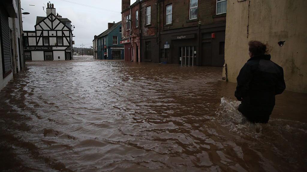 A woman wades through knee-deep water towards sandstone properties and a black and white clad pub near to the Whitesands in Dumfries