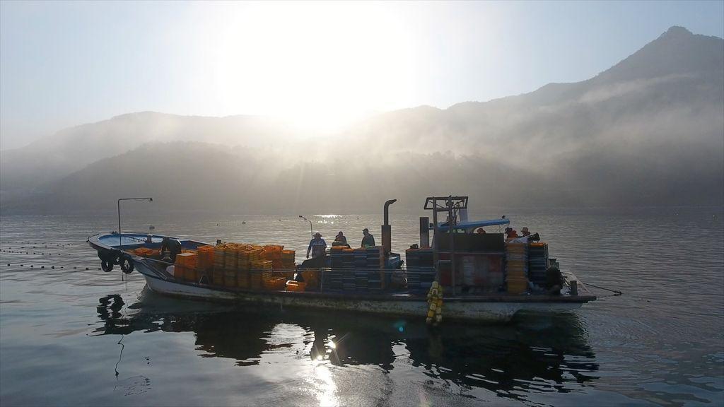 A small fishing board with people on it is seen in the water with mist and mountains behind.