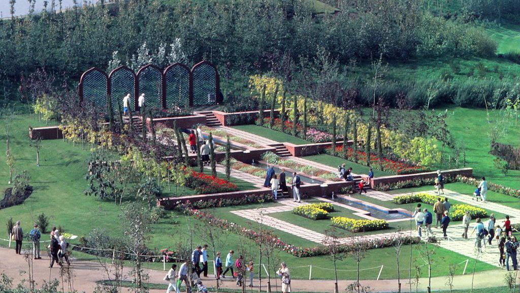 People walk round flower beds on part of the exhibition site. Pathways and colourful flower beds can be seen across a green lawn.