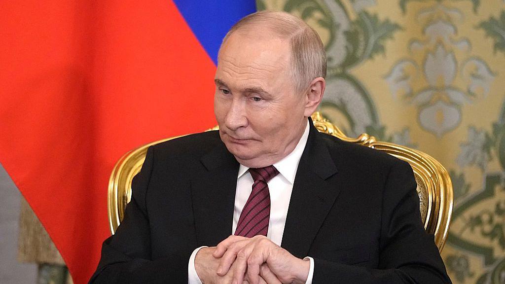 Vladimir Putin sitting in a gold chair, hands clasped and looking down. The Russian flag is draped behind him.