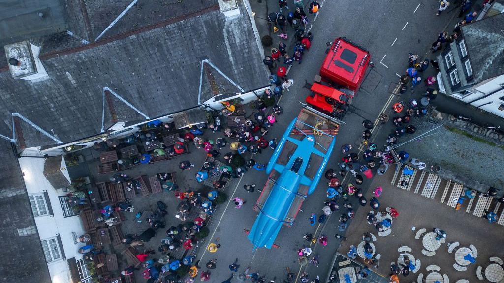 Aerial image showing Bluebird fastened to a trailer as it is transported through Coniston in March 2024. Crowds of people are lining the village's streets.