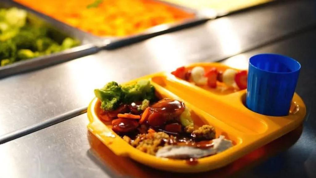 A yellow school tray of food with separate sections filled with chicken, broccoli and gravy and a blue beaker.