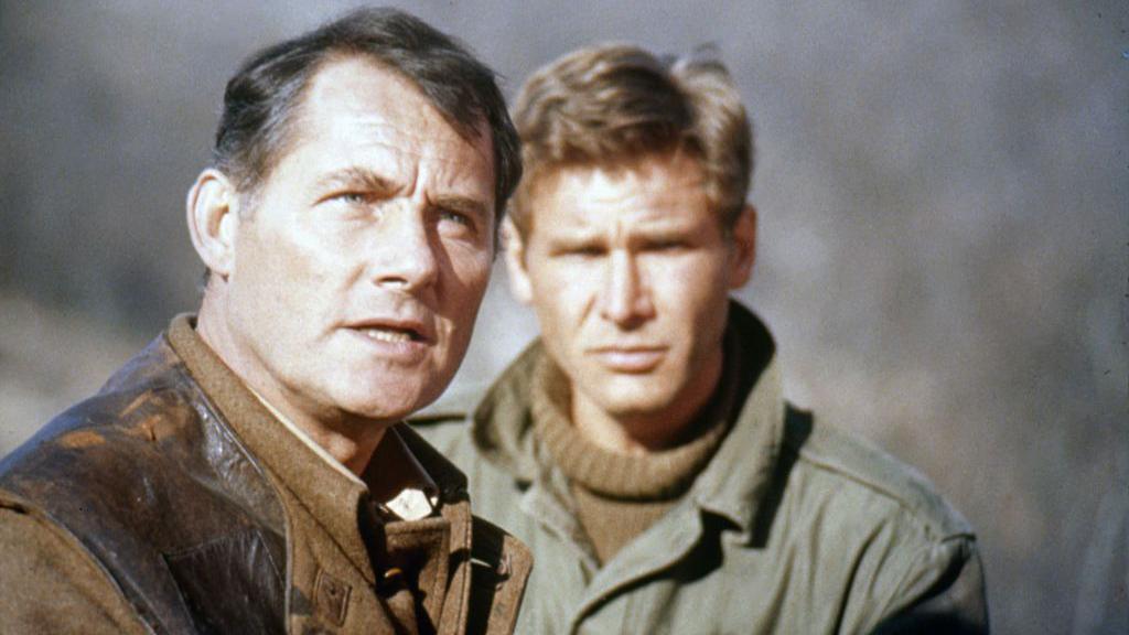 Robert Shaw in brown military jacket is standing on the left. Harrison Ford, in green military jacket, is to his right and slightly further back. They are looking towards the distance and the background is entirely out of focus.