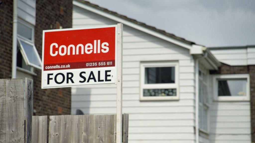 Connells for sale sign - written in white letters on a red background - attached to a wooden fence. A house with white wooden cladding can be seen in the background.
