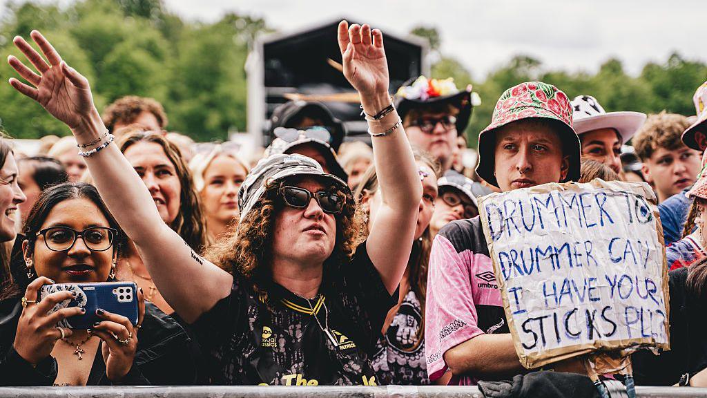 Attendees watch The Sherlocks performance during Tramlines at Hillsborough Park. One festival goer holds up a sign asking for drum sticks.