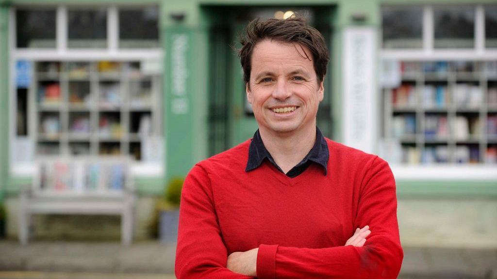 Adrian Turpin, artistic director of the Wigtown Book Festival, outside a bookshop in Wigtown. He has brown hair and is wearing a red t-shirt.