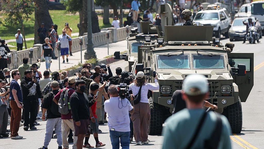 Protesters film in the streets as an armoured vehicle carrying federal troops drives next to LA's MacArthur Park 