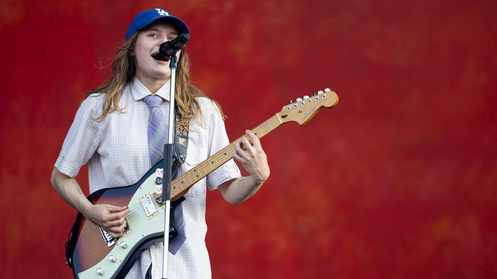 Music artist Girl In Red is on a stage with a red backdrop. She has brown hair. She is wearing a blue baseball cap, a white shirt, and a blue tie, She is holding a guitar, and singing into a microphone which is on a stand