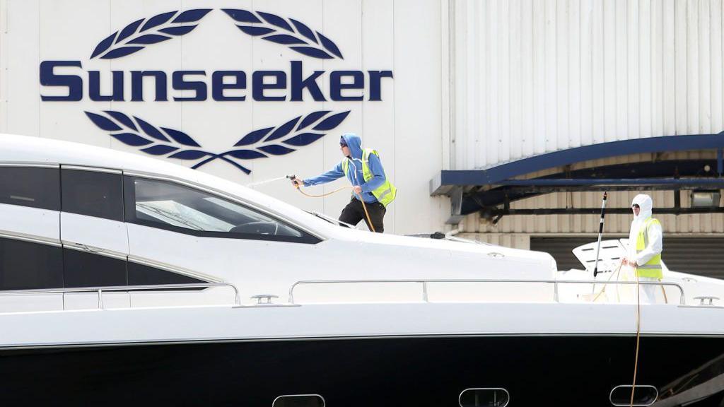 Two people using hoses to wash a luxury yacht, with a building behind them with the Sunseeker logo on it.