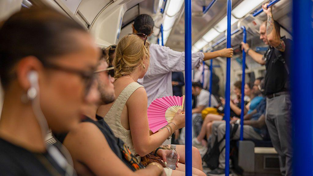 A London Underground carriage filled with people sitting down and two people are standing. The one person in focus is a woman with blonde hair facing away from the camera. She has a pink fan held in her hand and sunglasses on her head.