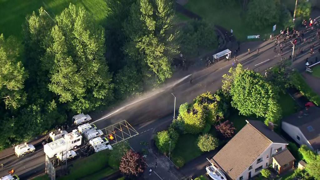 An aerial view of the water cannon in action. It is pushing back people who have gathered on the road. There are trees and houses on either side of the road.