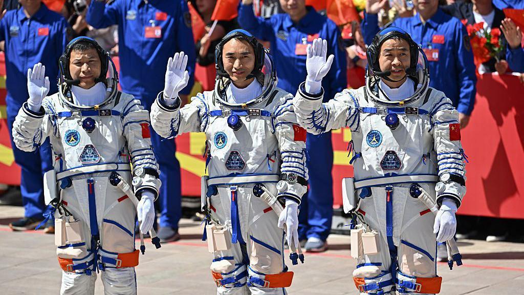 Wang Jie, Chen Zhongrui, and Chen Dong stand in space suits waving at a crowd of people.