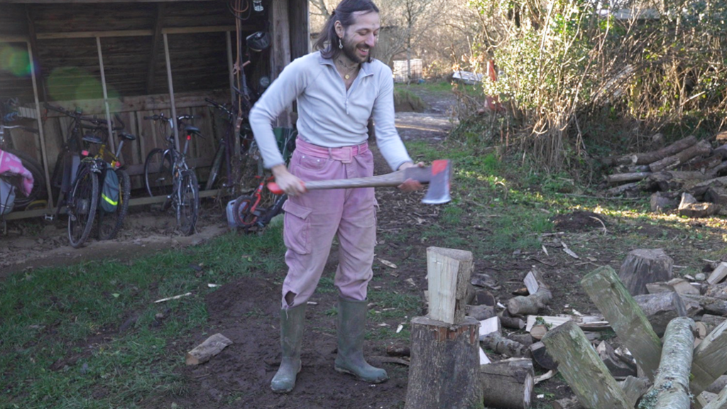 Andromeda chopping wood. Andromeda has shoulder-length dark hair and a short beard, a tattoo of a crescent moon on their head and is wearing drop earrings, two necklaces, pink trousers, wellies and a long sleeved white t-shirt. In front of them is a pile of wood and behind is a wooden bike sheds with several bikes in it.