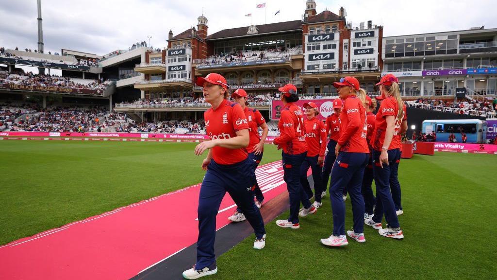 England women cricket players in red jerseys and caps walk across the field in front of a packed stadium, with the historic pavilion building visible in the background.