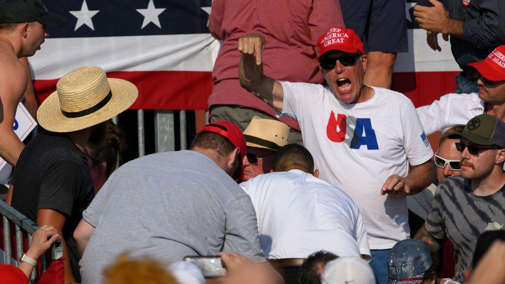 A man looks to be shouting and pointing down at a group of people huddled in a crowd, a scene of panic after shots were fired