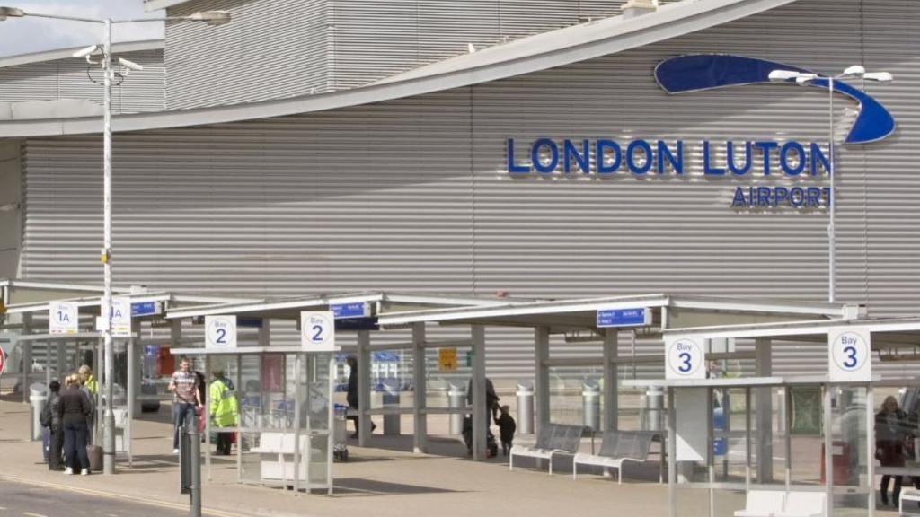 The exterior of London Luton Airport. The branding on the airport building is visible, in blue, and in the foreground are bus stands and some people standing in the sunshine.