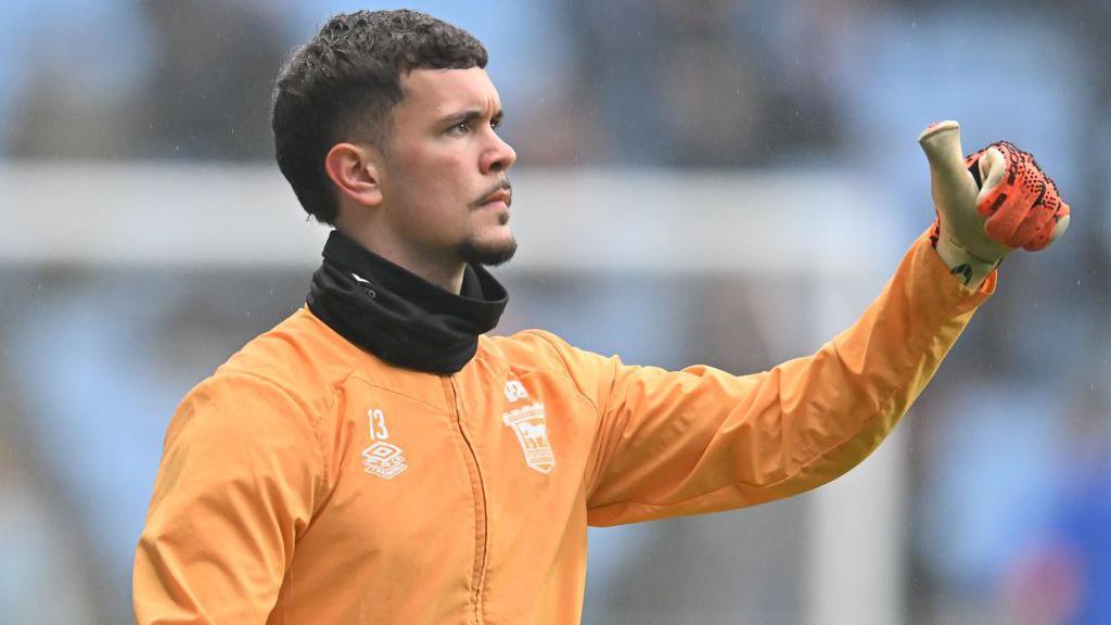 Cieran Slicker gives a thumbs up while wearing an orange Ipswich Town training jacket and goalkeeper gloves