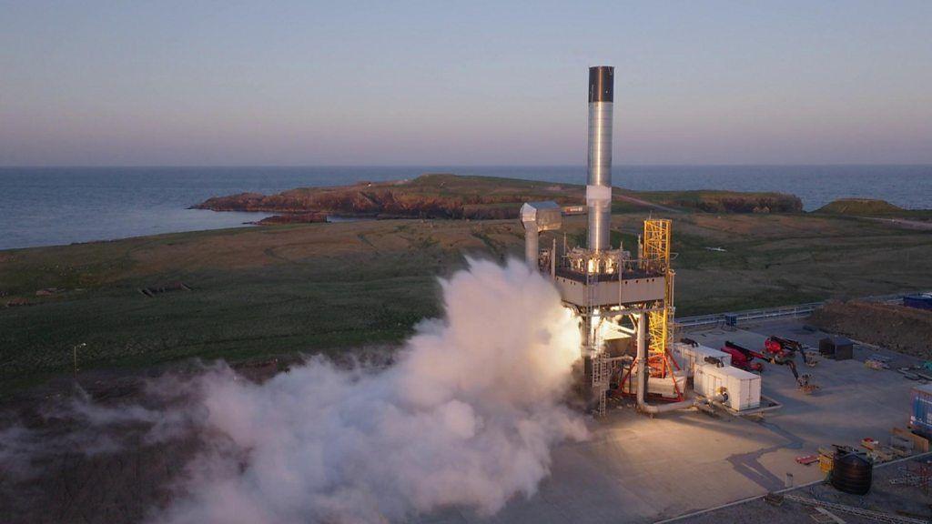 A cylindirical silver rocket without a nose cone on the launch pad at a peninsula on an island in Shetland. White vapour is emerging from its thrusters as it prepares for launch. In the background is the sea.